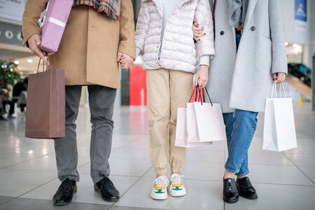 Lower part of casual family of three holding paperbags with purchases while spending time in the...