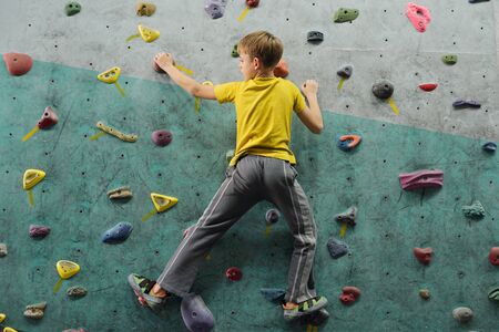 Back view of active schoolboy in sportswear moving upwards along climbing wall