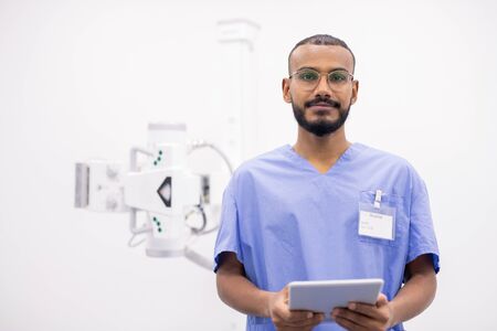 Young bearded mixed-race intern in uniform and eyeglasses using touchpad