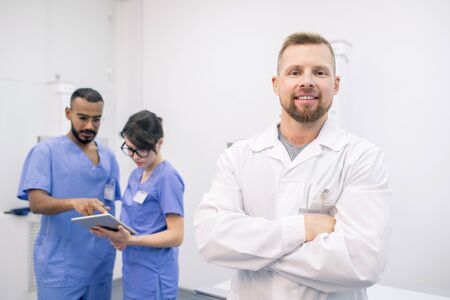 Young successful bearded clinician in whitecoat standing in front of camera