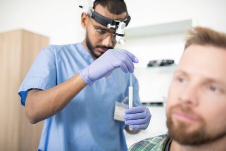 Young serious doctor in gloves putting medical stick into flask before treatmentの写真素材