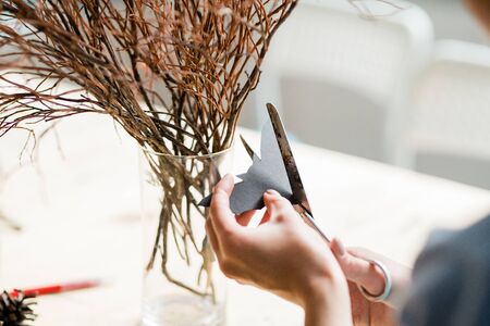 Hands of woman cutting paper bat with scissors to decorate dry branches in glass
