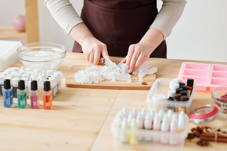Hands of craftswoman cutting transparent soap mass on board by wooden table before further process...