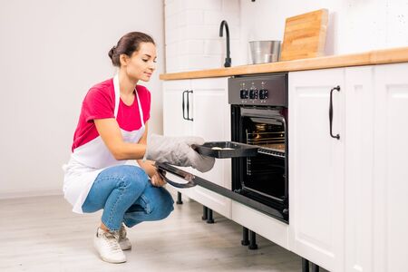 Young housewife squatting while putting tray with raw cookies into electric ovenの写真素材