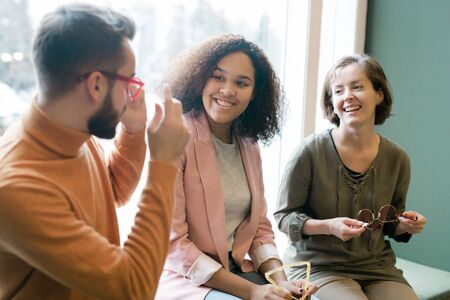 Two young cheerful multicultural women looking at man in new eyeglassesの写真素材