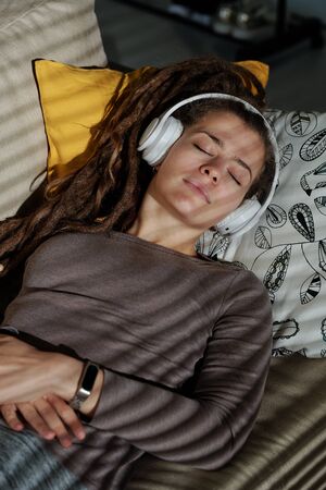 Pretty young relaxed woman with headphones lying on soft couch in dark roomの写真素材