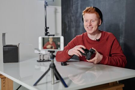 Young cheerful man in casualwear presenting his new photocamera in studio