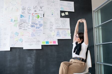 Young tired businesswoman stretching arms while sitting on chair by blackboard