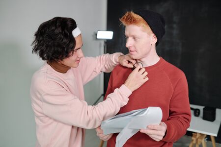 Casual guy putting microphone on sweatshirt of young male vlogger with paper