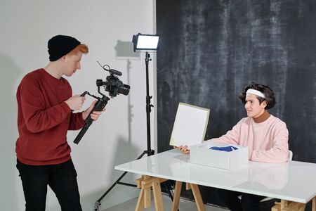 Serious young man with video shooting equipment standing in front of his friend