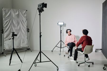 Two young male vloggers in casualwear sitting on chairs in front of each other in studio and...