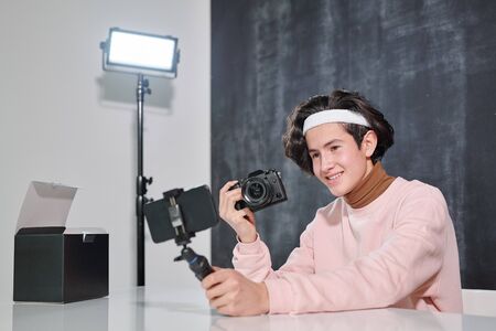 Young smiling male vlogger with photocamera sitting by desk and shooting himself
