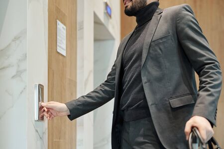 Young elegant businessman pushing button on the wall while standing by door and waiting for elevator...