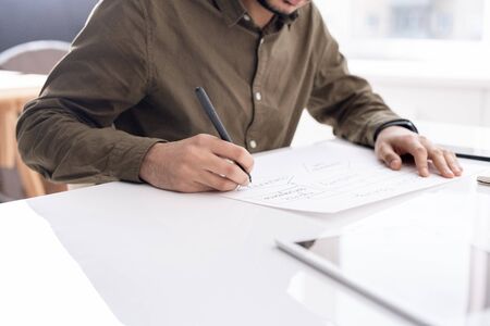Young busy economist drawing flow chart on paper while sitting by desk in office and preparing...