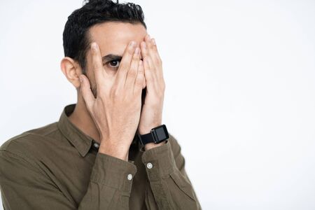 Young male employee covering his face by hands and looking at you by one eyeの写真素材