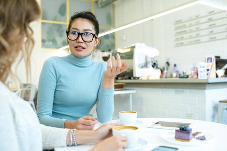 One of young women explaining something to her friend by cup of cappuccinoの写真素材