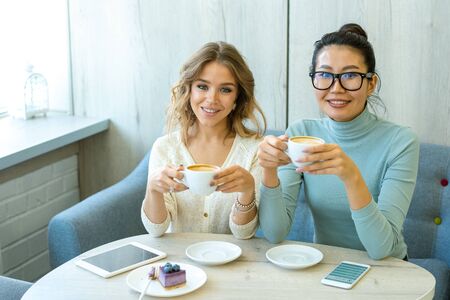 Cheerful girls in casualwear holding cups with coffee over table in cozy cafe