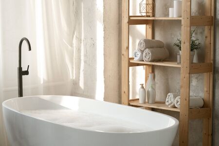 Large porcelain white bathtub filled with water and foam by wooden shelves