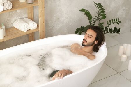 Young relaxed man with closed eyes lying in bathtub with hot water and foamの写真素材