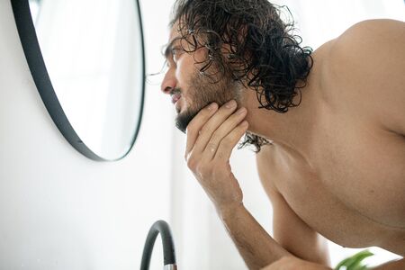 Shirtless guy bending over sink in front of mirror while touching his beard