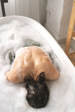Young naked man keeping his face in water while sitting in bath with foam