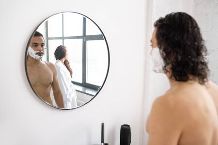 Shirtless young man with shaving foam on his beard standing in front of mirrorの写真素材