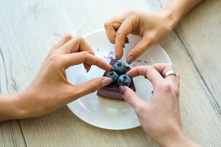 Hands of two girls taking blueberries from top of tasty cheesecake on plate while eating one cake together in cafeの写真素材