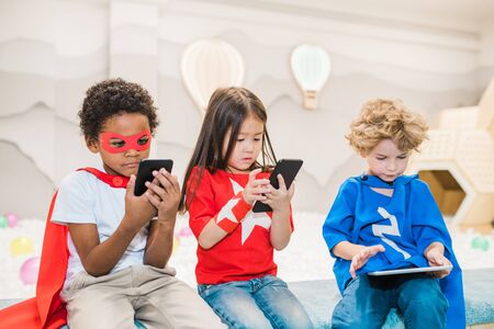 Cute little intercultural friends with mobile gadgets playing games at leisureの写真素材