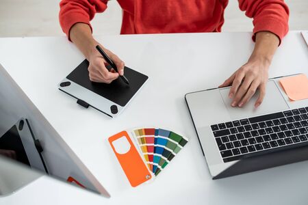 Hands of young web designer holding stylus over screen of graphics tablet while using laptop during workの写真素材