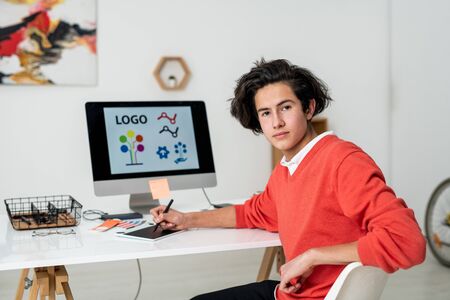 Young serious web designer with stylus and graphics tablet sitting by desk