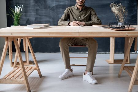 Young casual student sitting by wooden table while writing essay or carrying out home assignment in notepadの写真素材