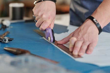 Close-up of unrecognizable man using cutting wheel and metal ruler while producing leather goods at tableの写真素材