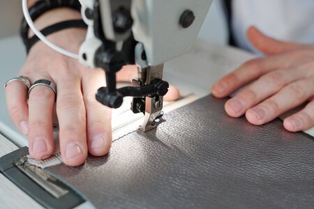Close-up of unrecognizable man with rings using sewing machine while stitching zipper and leather...