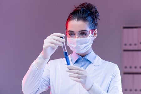 Young woman in mask, gloves and eyeglasses looking at blue substance in flaskの写真素材