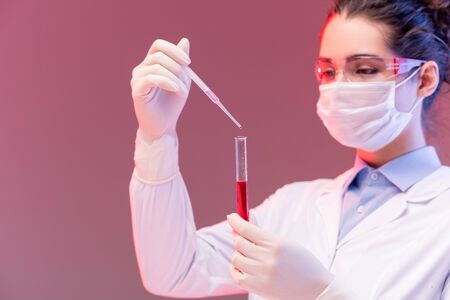 Young chemist in protective eyeglasses and mask dropping red fluid into flask