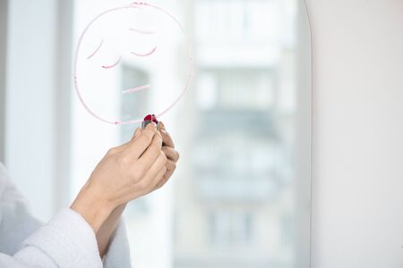 Hand of young female in bathrobe with lipstick drawing upset face on mirror while expressing her despairの写真素材
