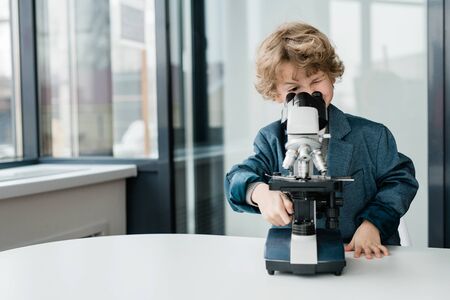 Clever cute schoolboy or biologist standing by desk while looking in microscopeの写真素材