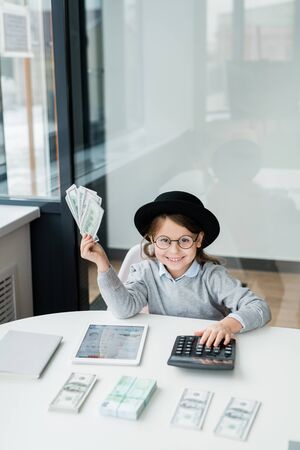 Little cheerful accountant in hat and casualwear counting your salary by deskの写真素材