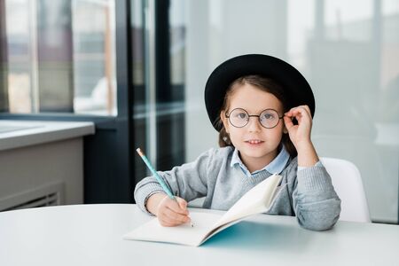 Adorable clever schoolgirl in hat looking at you while making notes in copybook