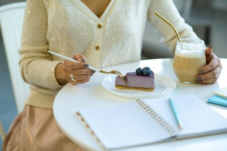 Hands of young casual woman taking piece of tasty blueberry cheesecake and having cappuccino while sitting in cafeの写真素材
