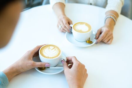 Hands of two friendly girls sitting by table in cafe and having fresh tasty cappuccino during...