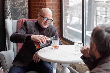 Smiling bald man in glasses sitting at table and pouring whisky in glass while meeting with business partner in restaurantの写真素材