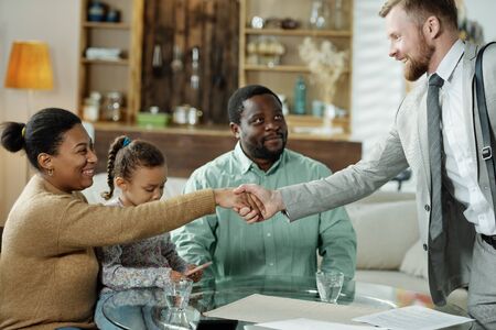 Side view of cheerful handsome adviser shaking hand of woman while meeting with ethnic family to give advise on home loanの写真素材