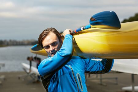 Athlete with his watercraft. Young Caucasian blond-haired man carrying yellow kayak and paddle after training on waterの写真素材