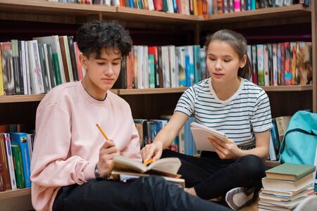 Clever confident girl pointing at page of book held by her classmate