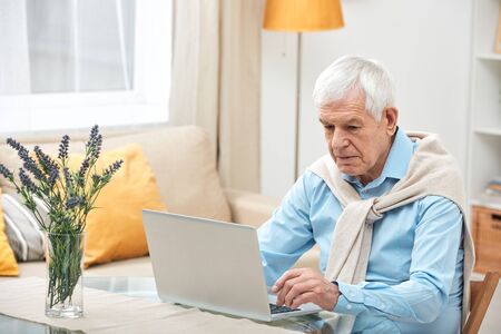 Senior man in casualwear sitting by table in front of laptop while surfing in the net and looking...