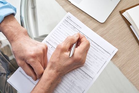 Hands of contemporary senior retired man with pen filling in health insurance form while sitting by tableの写真素材
