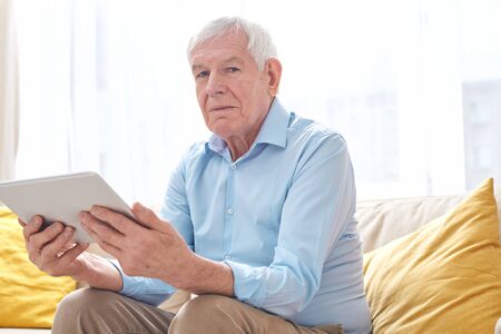 Serious senior man in casualwear looking at you while sitting on couch and using digital tablet in...