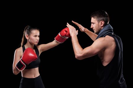 Young serious fit woman in activewear and boxing gloves hitting trainer