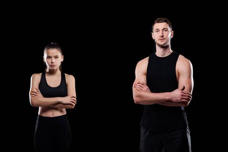 Serious young sporty couple in activewear crossing arms by chest while standing in front of camera on black backgroundの写真素材